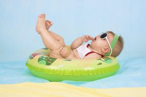 Baby mit weißer Sonnenbrille und grünem Haarband liegt entspannt auf einem grünen Rettungsring Und treibt auf blauem Wasser